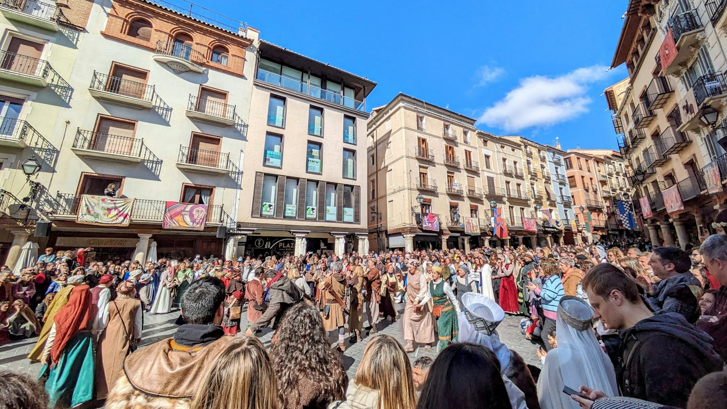 Menschen in historischen Kostümen auf einem sonnigen Stadtplatz mit Bannern.